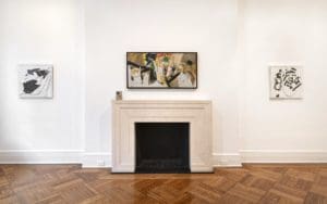 The wall in a gallery with parquet wood flooring featuring a painting over a fireplace.